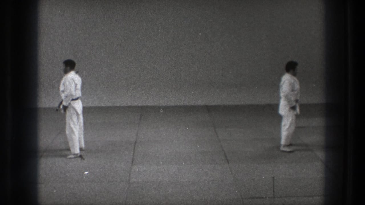 Two martial artists in white gi stand facing each other on a plain indoor training floor.