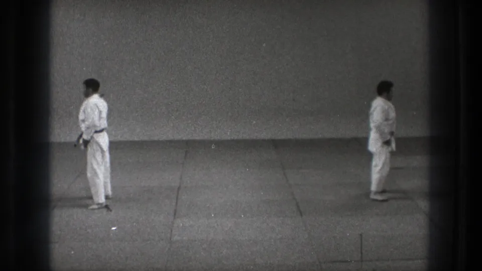 In Tokyo 1971 footage, two martial artists in white gi face each other in a formal stance on a plain dojo-like floor.