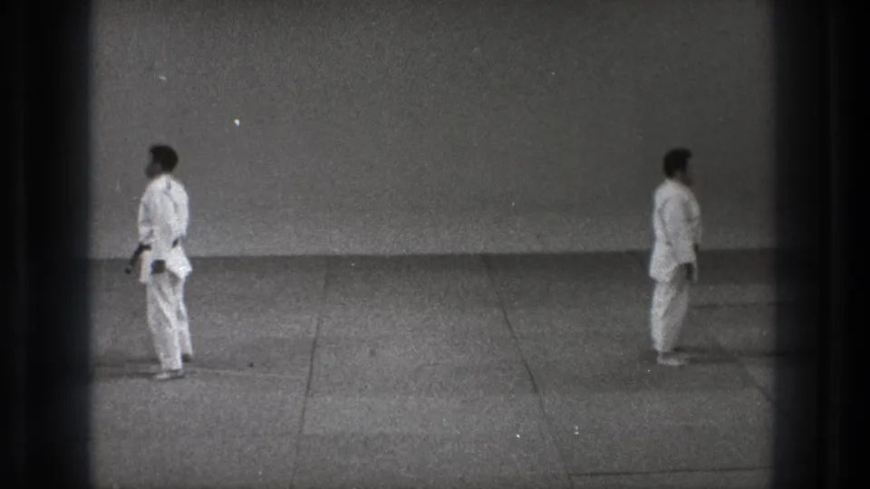Two martial artists in white gi stand separated in a formal stance, with visible 8mm frame edges and grain.