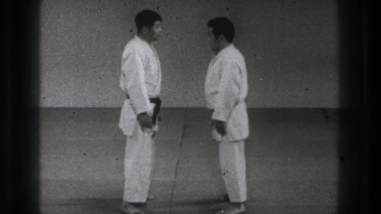 Two adult martial artists in white uniforms and dark belts stand facing each other in Tokyo home-movie footage from 1971.