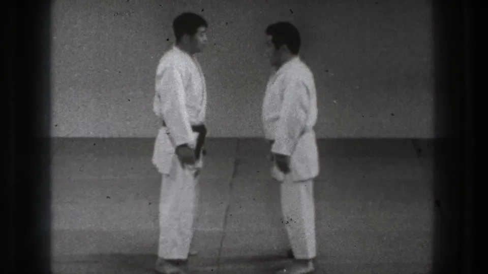 Two adult martial artists in white uniforms and dark belts stand facing each other in Tokyo home-movie footage from 1971.