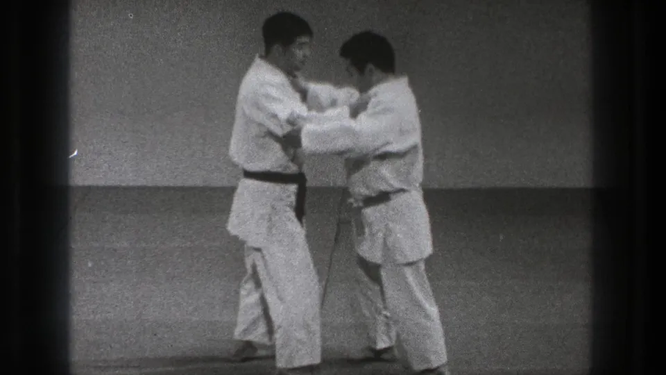 Two adult martial artists in white uniforms begin close-contact grappling on a plain indoor mat in 1971 Tokyo footage.