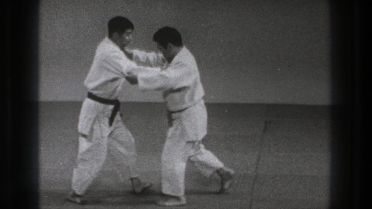 Two adult martial artists in white uniforms hold each other's jackets while shifting foot positions on the mat.