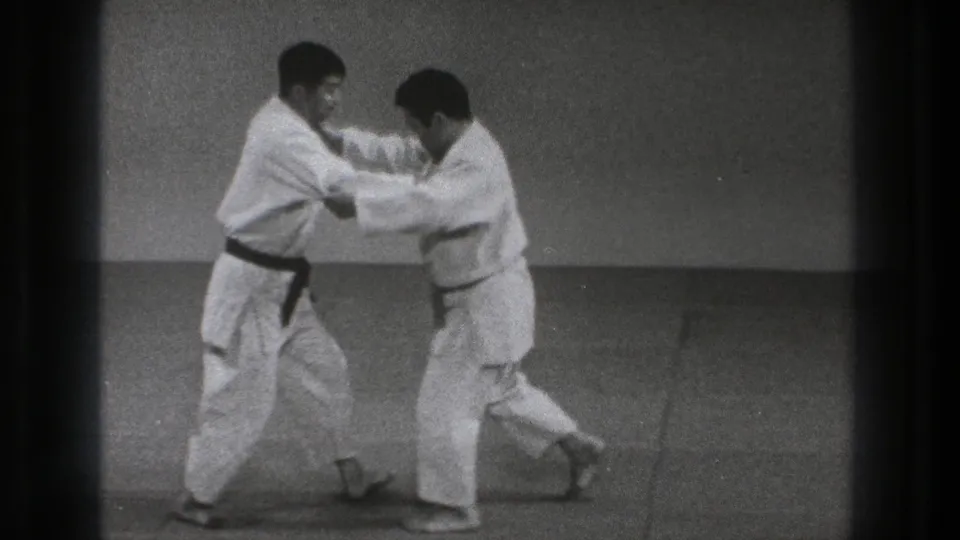 Two adult martial artists in white uniforms hold each other's jackets while shifting foot positions on the mat.