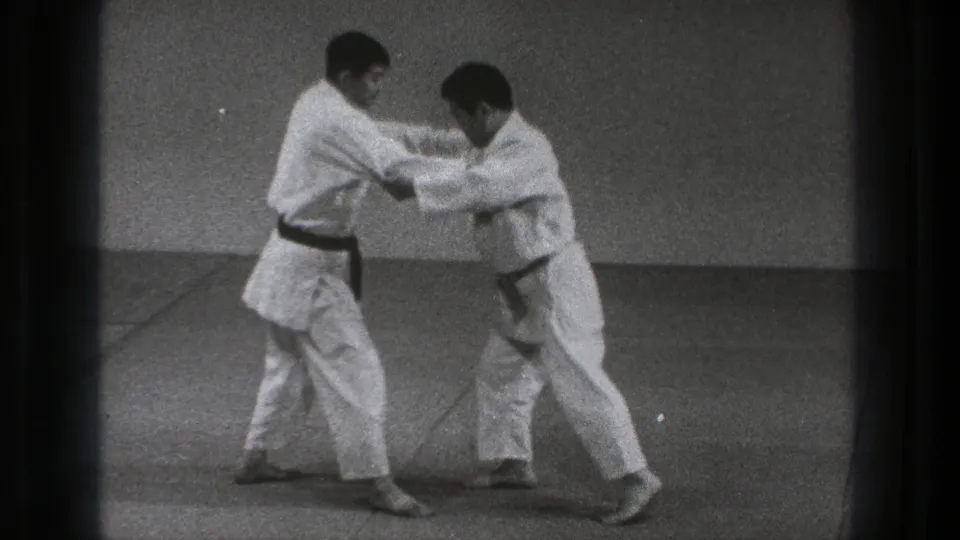 Two adult martial artists in 1971 Tokyo footage lean into a close grip with bent knees and braced footing on the mat.