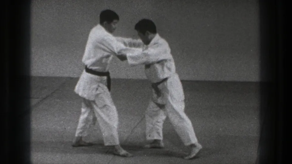 In Tokyo, Japan in 1971, two karate practitioners in white gi hold each other at the arms on a simple mat.