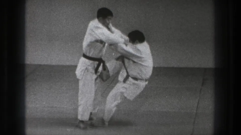 The black-belt practitioner begins the throw, drawing his partner off balance on a plain indoor mat in 1971 Tokyo footage.