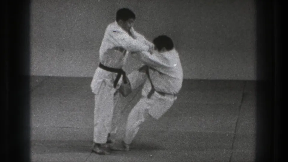 During the judo-style technique, one man lowers toward the mat while the standing practitioner controls the grip and balance.