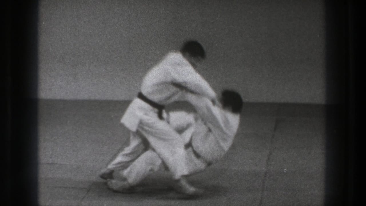 In a 1971 Tokyo indoor practice scene, two adult men in white gi engage in the opening movement of a self-defense throw.