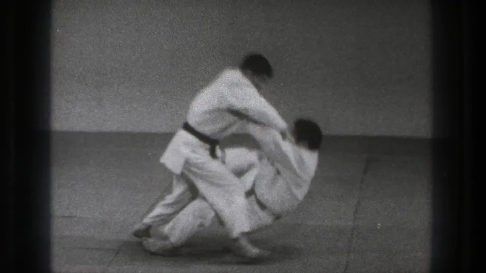 In a 1971 Tokyo indoor practice scene, two adult men in white gi engage in the opening movement of a self-defense throw.