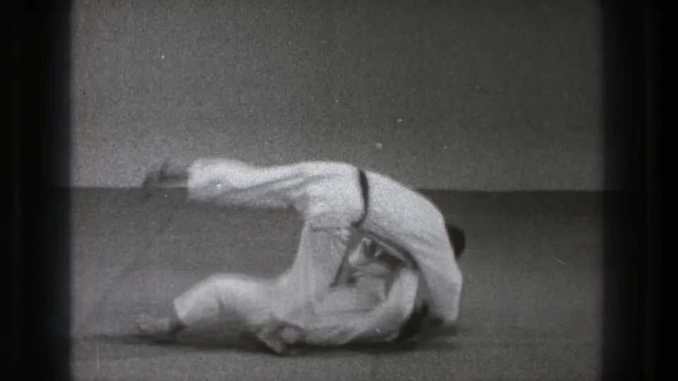 A white-uniformed practitioner rolls through a throw as his partner flips over him on the mat in the Tokyo training footage.