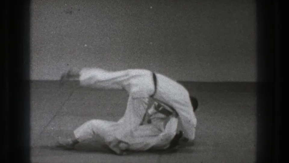 The martial arts pair remains low to the padded floor as the throw completes in grainy black-and-white 8mm footage.