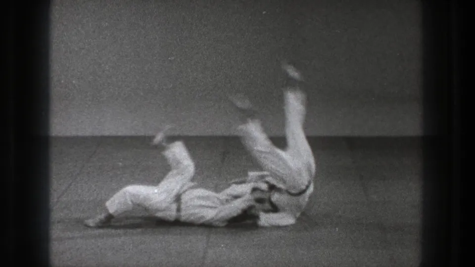 Tokyo 1971 8mm frame of two adult judo practitioners in white gi during a throw on a plain dojo mat.