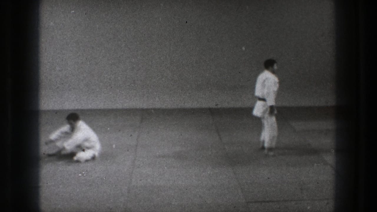 Tokyo 1971 8mm frame of a judo practice reset, with one adult seated on the mat and another standing nearby.