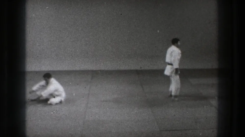 Tokyo 1971 8mm frame of a judo practice reset, with one adult seated on the mat and another standing nearby.