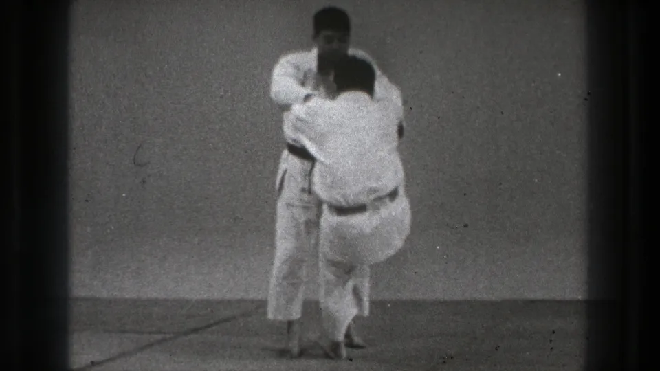 In supported Tokyo 1971 8mm footage, two men in white gi begin a close-contact karate practice movement on a plain mat.