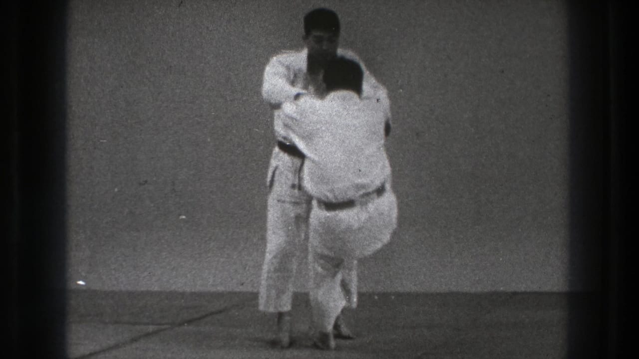 Two men in white gi maintain close grips during a controlled martial arts throw practice in the Tokyo 1971 home-movie clip.