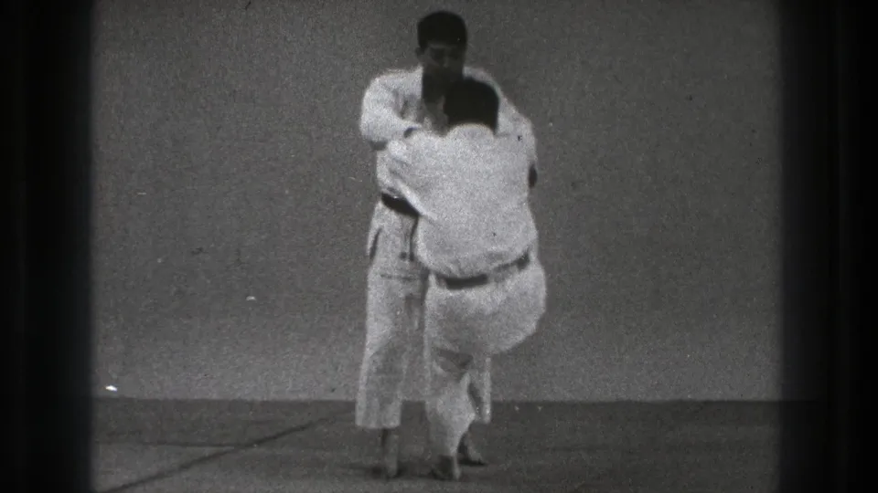 Two men in white gi maintain close grips during a controlled martial arts throw practice in the Tokyo 1971 home-movie clip.