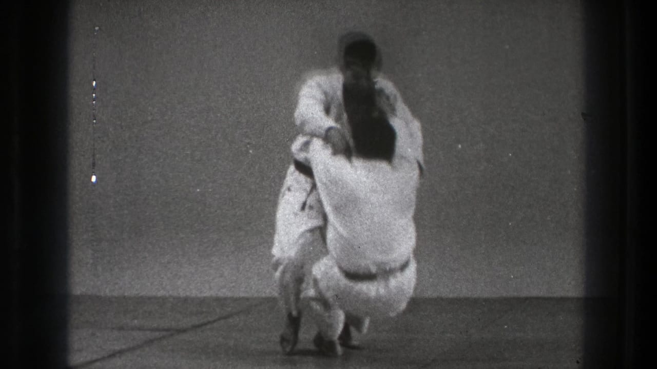 A man in a white gi drops into a lower stance as his partner remains upright during the supported Tokyo 1971 martial arts practice.