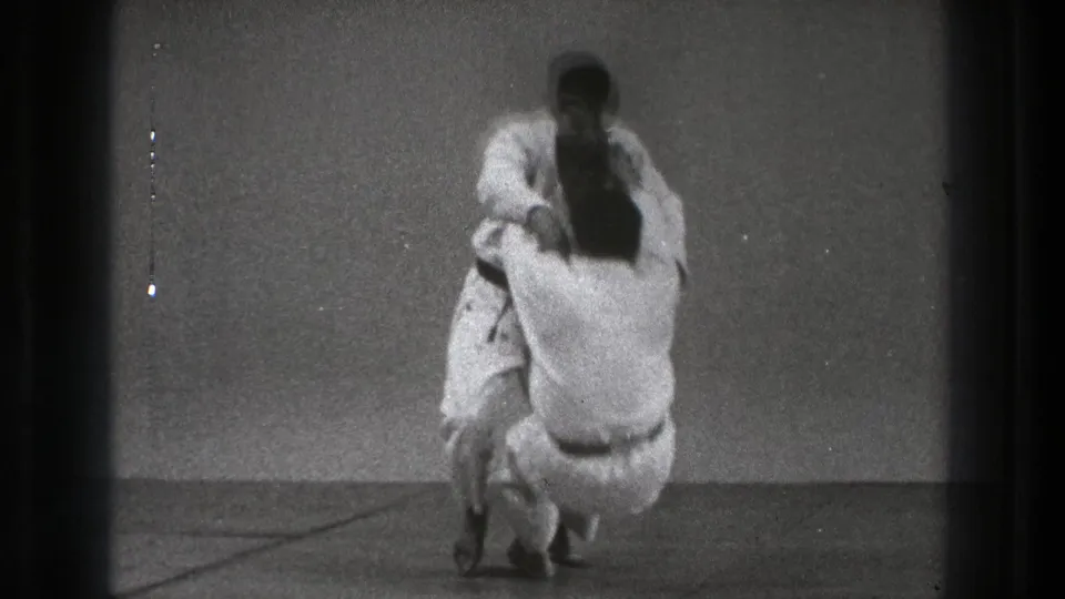 A man in a white gi drops into a lower stance as his partner remains upright during the supported Tokyo 1971 martial arts practice.