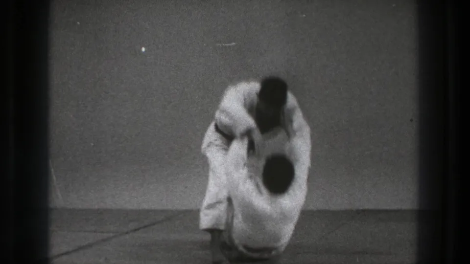 In 1971 Tokyo footage, two adult judo practitioners in white gi and black belts grapple on a plain indoor mat.