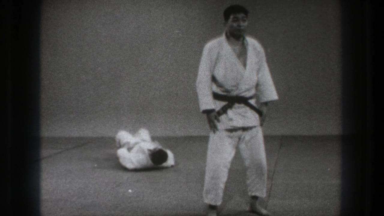 After the throw, one adult practitioner stands in a white gi and black belt while his partner lies curled on the mat behind him.