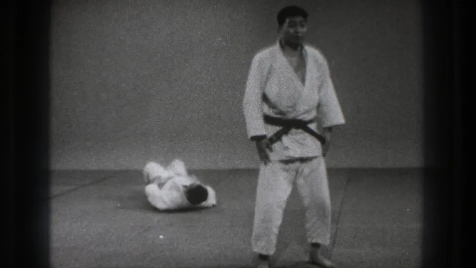 After the throw, one adult practitioner stands in a white gi and black belt while his partner lies curled on the mat behind him.