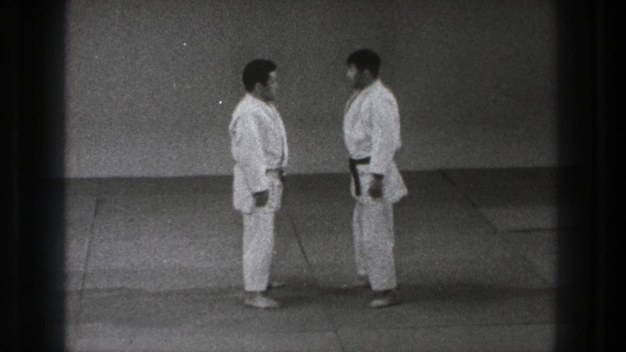 The two adult judo practitioners stand face to face in white gi and black belts on a plain indoor mat in Tokyo footage from 1971.