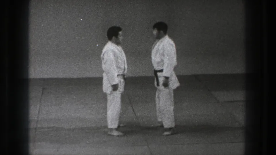 The two adult judo practitioners stand face to face in white gi and black belts on a plain indoor mat in Tokyo footage from 1971.