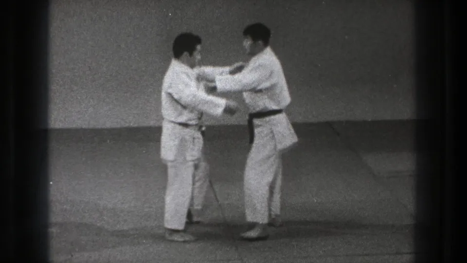 In 1971 Tokyo footage, two adult men in white gi stand on padded mats preparing a close-contact karate or judo-style technique.