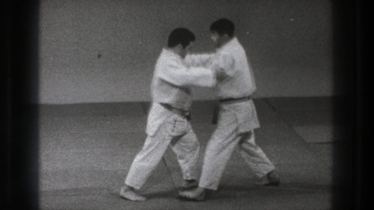 The two adult practitioners in white gi move into a close grip on the dojo mat, with one wearing a dark belt and the other a lighter belt.