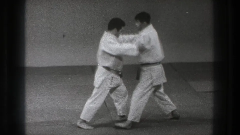 The two adult practitioners in white gi move into a close grip on the dojo mat, with one wearing a dark belt and the other a lighter belt.