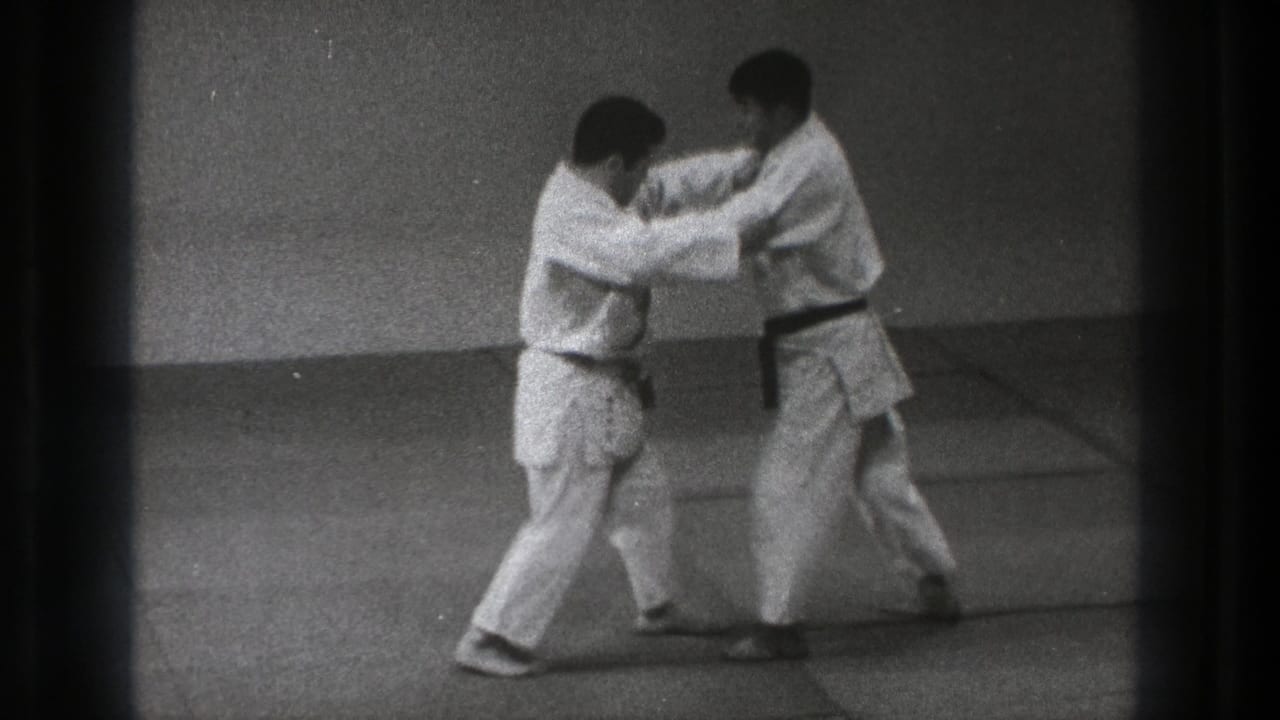 In the black-and-white 8mm frame, two adult men in gi lean forward while practicing a close-contact technique on padded dojo mats.