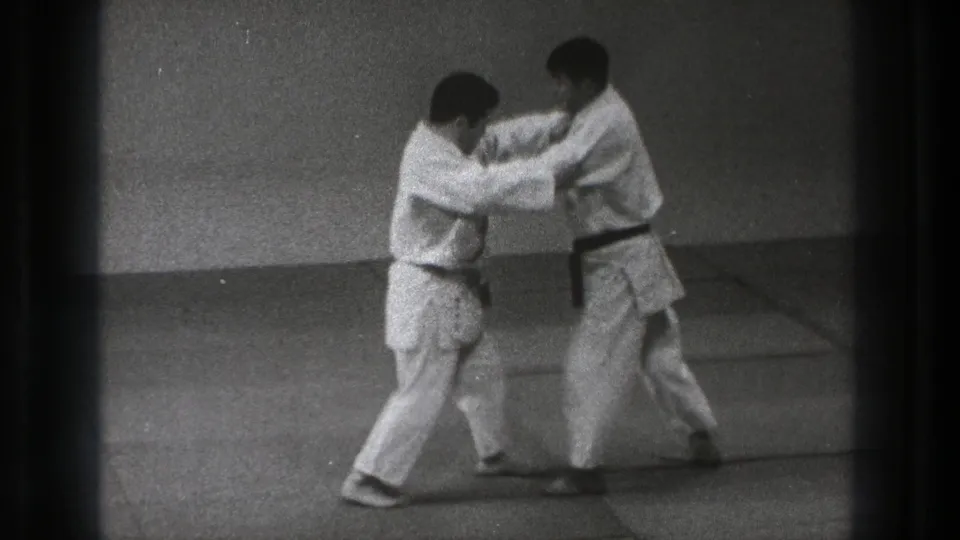 In the black-and-white 8mm frame, two adult men in gi lean forward while practicing a close-contact technique on padded dojo mats.