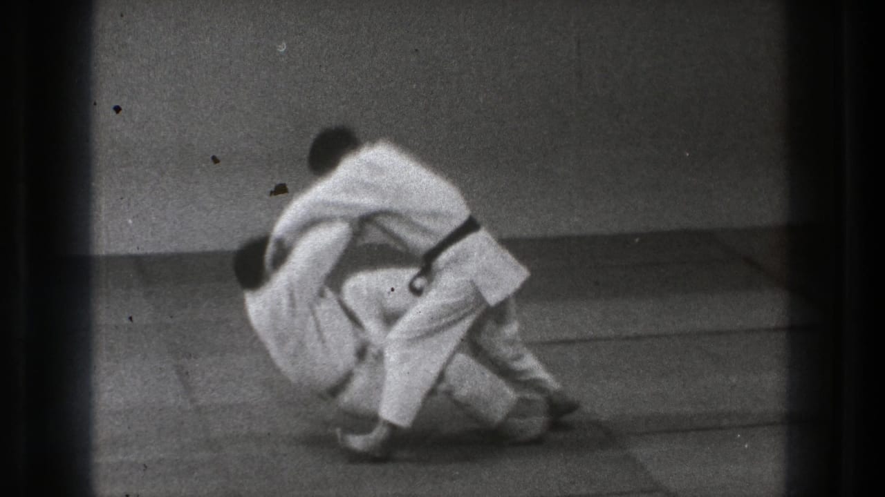 The 1971 Tokyo judo sequence reaches a controlled takedown, with one practitioner lowering his partner toward the mat.