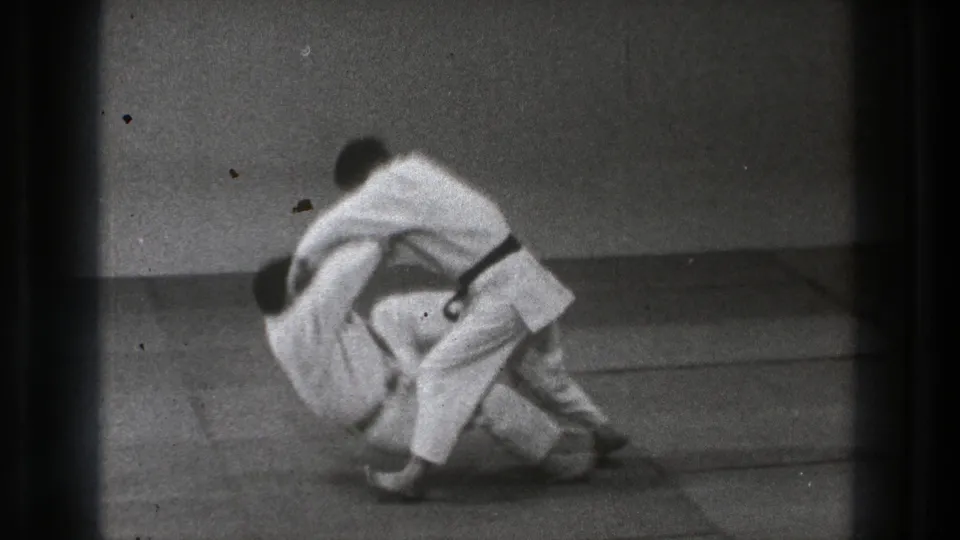 The 1971 Tokyo judo sequence reaches a controlled takedown, with one practitioner lowering his partner toward the mat.