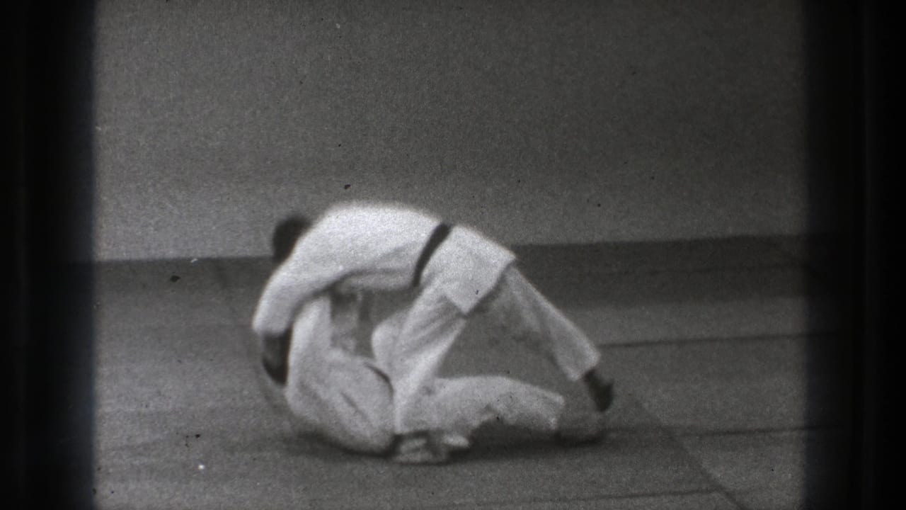 Two martial artists in white gi roll low on a padded mat.