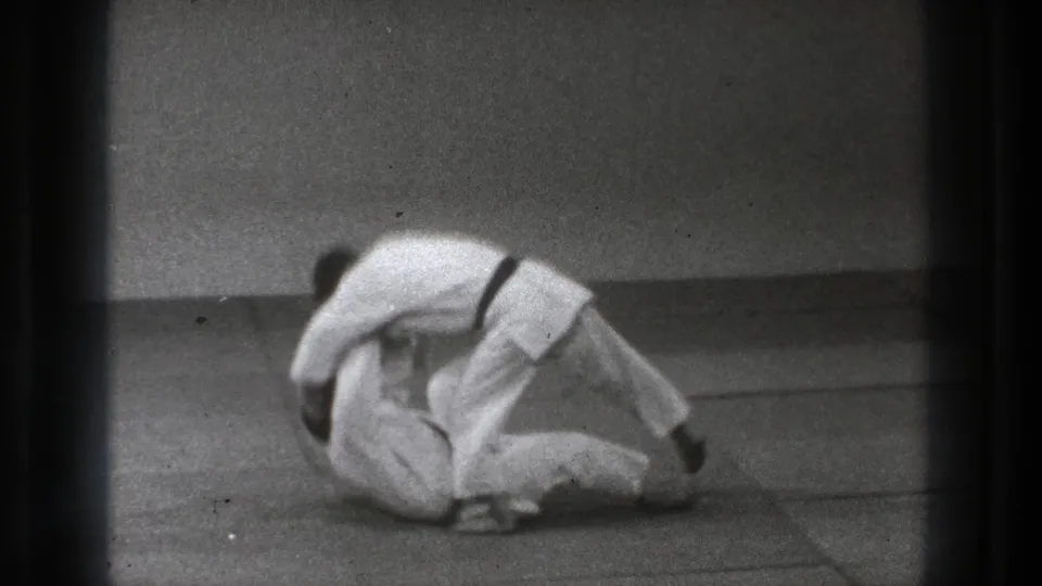 In 1971 Tokyo footage, two martial artists in white gi are low on the mat during the start of a controlled throw or roll.