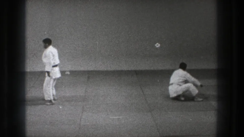 At the end of the demonstration, one practitioner stands in a white gi while the other sits on the mat in the sparse dojo space.