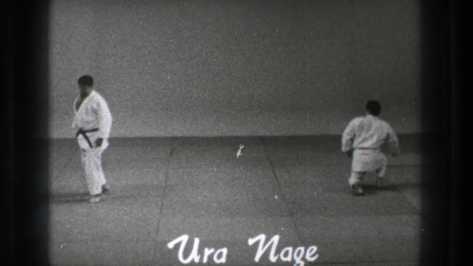 1971 Tokyo 8mm frame showing two men in white judogi on a plain mat, with one kneeling and Ura Nage written on screen.