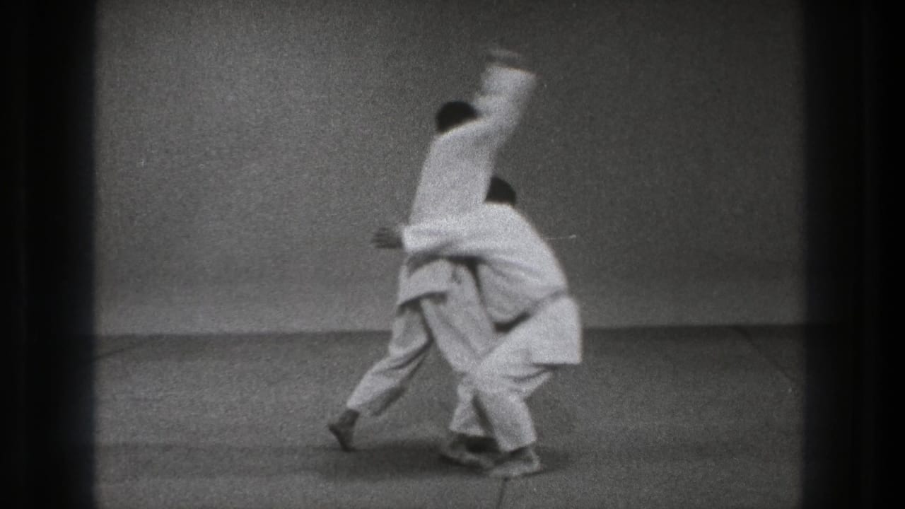 In 1971 Tokyo footage, two adult practitioners in white gi start an upper-body throwing technique inside a simple dojo-like room.