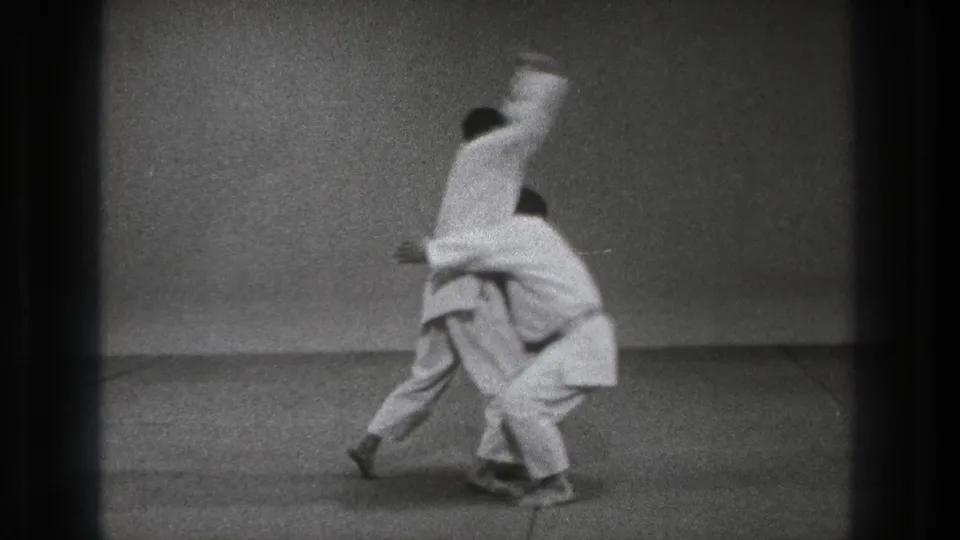 In 1971 Tokyo footage, two adult practitioners in white gi start an upper-body throwing technique inside a simple dojo-like room.