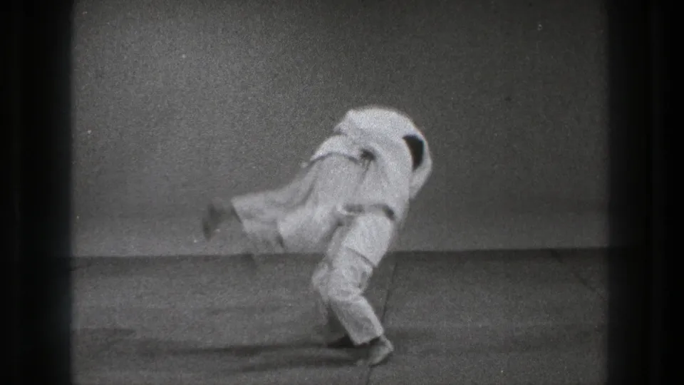 A gi-clad martial arts practitioner lifts his partner off balance during a dynamic throw in the 1971 Tokyo indoor practice footage.