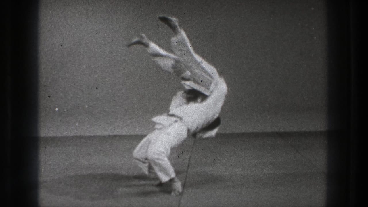 The thrown practitioner is nearly inverted with legs raised while his partner completes the motion on the indoor practice floor.