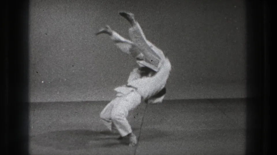 The thrown practitioner is nearly inverted with legs raised while his partner completes the motion on the indoor practice floor.