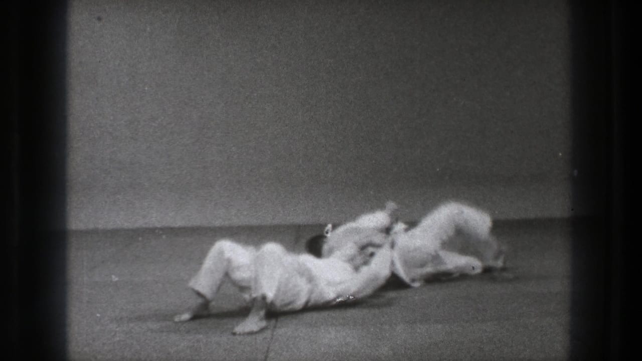 Two adult men in white gi practice a floor grappling move on a mat in 1971 Tokyo footage.