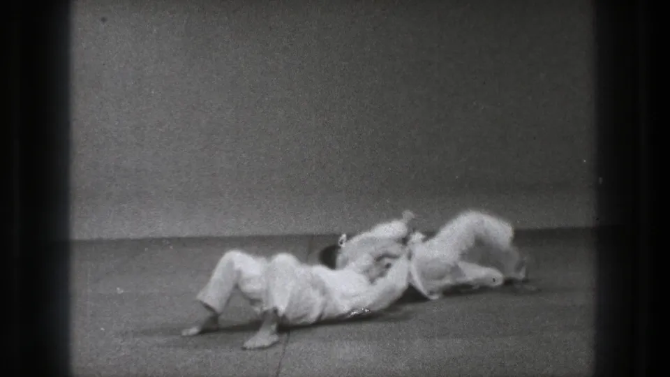 Two adult men in white gi practice a floor grappling move on a mat in 1971 Tokyo footage.