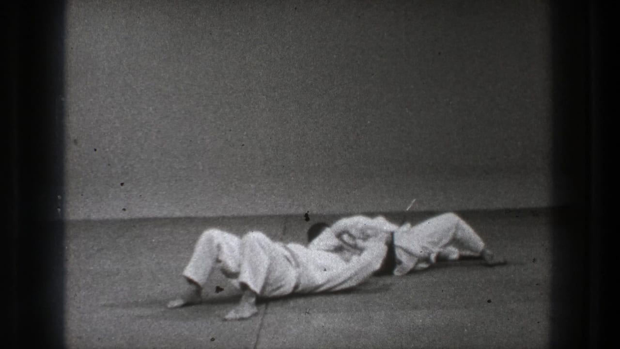 Two adult martial-arts practitioners are on the mat in a close hold during the Tokyo training sequence.