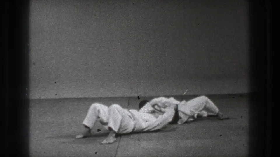 Two adult martial-arts practitioners are on the mat in a close hold during the Tokyo training sequence.