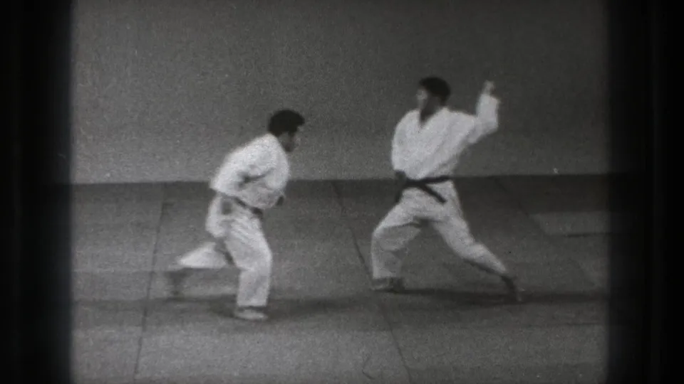 Two adult men in white gi stand facing each other on a practice mat in the 1971 Tokyo clip.